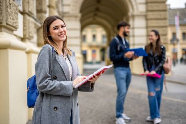 Neşeli bir kız öğrenci ön planda duruyor, bir yığın kitap tutarken kameraya sıcak bir şekilde gülümsüyor. Onun arkasında, iki sınıf arkadaşı görünür, biraz bulanık, işbirlikçi bir akademik ortam öneriyor.. 