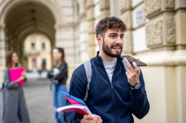 Odaklanmış bir öğrenci elinde kitaplarla ön planda duruyor. Akıllı telefonunu kullanarak sesli mesaj gönderiyor.. 