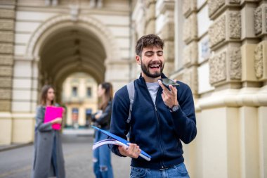 Odaklanmış bir öğrenci elinde kitaplarla ön planda duruyor. Akıllı telefonunu kullanarak sesli mesaj gönderiyor.. 