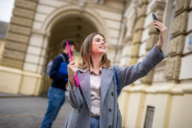 Gülümseyen bir öğrenci bir yığın kitap tutar ve akıllı telefonuyla bir selfie çeker, bir anlık neşe ve bağlantı yakalar..