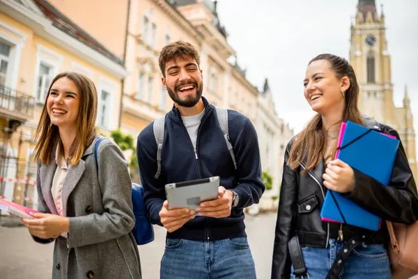 Üç öğrenci ellerinde kitap ve bir tablet tutuyor, eğitimsel içeriği tartışıyor ve paylaşıyorlar. Sırt çantasıyla gelişigüzel giyinmiş..