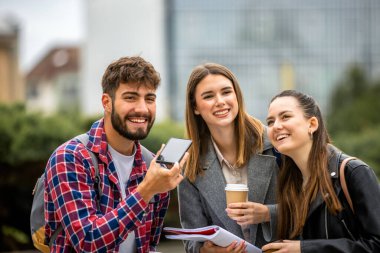 Üç öğrenci dışarıda toplanmış, akıllı telefondan bir sesli mesaj dinliyorlar.. 
