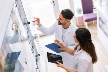 İki profesyonel eczacı eczane raflarının yanında durup, dijital tabletteki bilgileri dikkatle seçerek ilaç seçiyorlar.. 