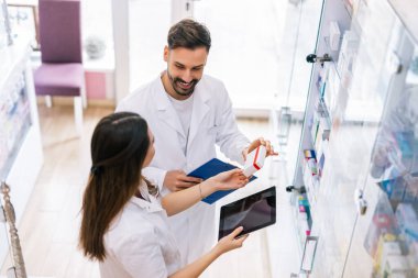 İki profesyonel eczacı eczane raflarının yanında durup, dijital tabletteki bilgileri dikkatle seçerek ilaç seçiyorlar.. 