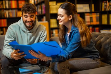 İki odaklanmış öğrenci sessiz bir kütüphane ortamında birlikte okuyorlar, bir klasörden içeriği okuyup tartışıyorlar.. 