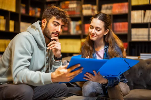 İki odaklanmış öğrenci sessiz bir kütüphane ortamında birlikte okuyorlar, bir klasörden içeriği okuyup tartışıyorlar.. 