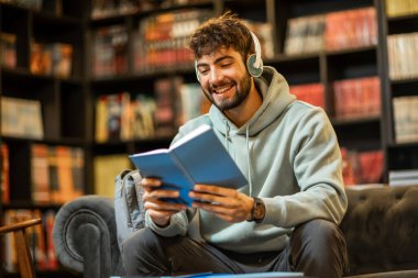 Bir erkek öğrenci sessiz bir kütüphanede oturuyor, kulaklık takıp fizik kitabı okuyor, sesli kitap ya da e-öğrenim materyali dinlediğini söylüyor..