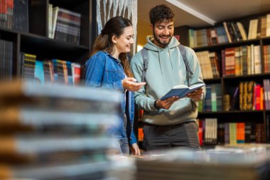 Genç bir erkek ve bayan öğrenci kütüphanedeki kitaplıklara göz atıyorlar..