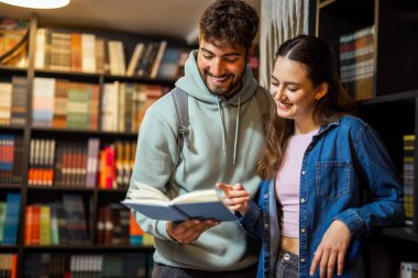 Genç erkek ve bayan öğrenci bir kütüphanede birlikte duruyorlar, aynı kitabı okuyorlar ve ortak çalışma anlarını paylaşıyorlar..