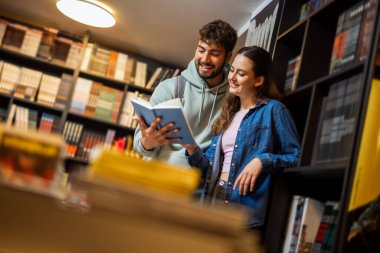 Genç erkek ve bayan öğrenci bir kütüphanede birlikte duruyorlar, aynı kitabı okuyorlar ve ortak çalışma anlarını paylaşıyorlar..