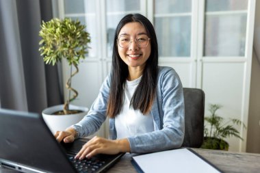 Taylandlı kadın rahat bir ev ortamında masa başında oturuyor, uzaktan bilgisayarına odaklanıyor..