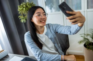Taylandlı kadın masasında rahat bir ofiste oturmuş bilgisayarında çalışırken akıllı telefonuyla selfie çekiyor.. 