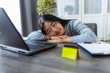 Bilgisayarında çalışan genç bir Taylandlı kadın son derece yorgun görünüyor ve başını masaya dayayıp yorgunluk ve yorgunluk belirtileri gösteriyor.. 