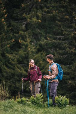 Sırt çantaları, yürüyüş direkleri ve açık hava kıyafetleriyle donatılmış genç bir erkek ve kadın, bereketli doğayla çevrili bir dağ yolu boyunca birlikte yürüyorlar. Çevrelerindeki manzaranın güzelliğine hayran olmak için dururlar..