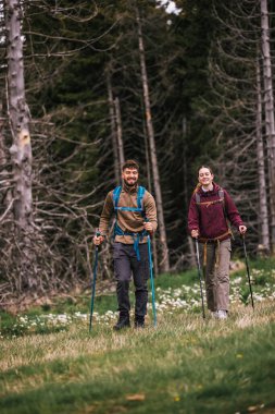 Sırt çantaları, yürüyüş direkleri ve açık hava kıyafetleriyle donatılmış genç bir erkek ve kadın, yemyeşil doğayla çevrili bir dağ yolunda birlikte yürüyorlar.