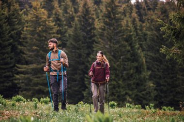 Sırt çantaları, yürüyüş direkleri ve açık hava kıyafetleriyle donatılmış genç bir erkek ve kadın, yemyeşil doğayla çevrili bir dağ yolunda birlikte yürüyorlar.. 