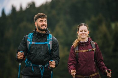 Sırt çantaları, yürüyüş direkleri ve açık hava kıyafetleriyle donatılmış genç bir erkek ve kadın, yemyeşil doğayla çevrili bir dağ yolunda birlikte yürüyorlar.