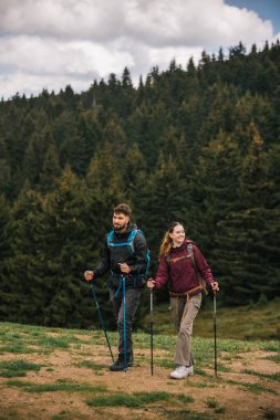 Sırt çantaları, yürüyüş direkleri ve açık hava kıyafetleriyle donatılmış genç bir erkek ve kadın, yemyeşil doğayla çevrili bir dağ yolunda birlikte yürüyorlar.
