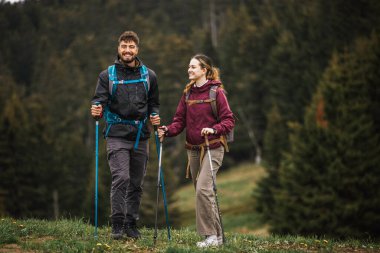 Sırt çantaları, yürüyüş direkleri ve açık hava kıyafetleriyle donatılmış genç bir erkek ve kadın, yemyeşil doğayla çevrili bir dağ yolunda birlikte yürüyorlar.