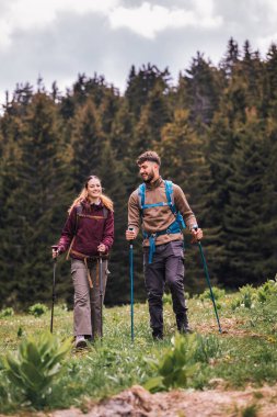 Sırt çantaları, yürüyüş direkleri ve açık hava kıyafetleriyle donatılmış genç bir erkek ve kadın, yemyeşil doğayla çevrili bir dağ yolunda birlikte yürüyorlar.. 