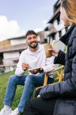 Genç bir adam ve bir kadın, hafta sonu kaçamağı sırasında şezlonglarda dinleniyorlar. Doğa ve temiz havayla çevrili olarak sıcak kahvenin ve ferahlatıcı meyve suyunun tadını çıkarırlar..