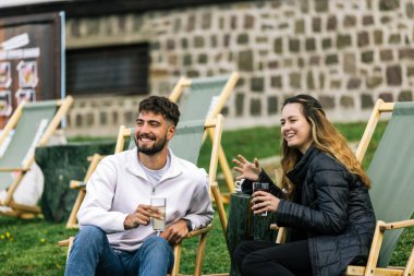 Genç bir adam ve bir kadın, hafta sonu kaçamağı sırasında şezlonglarda dinleniyorlar. Doğa ve temiz havayla çevrili olarak sıcak kahvenin ve ferahlatıcı meyve suyunun tadını çıkarırlar..
