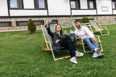 Genç bir erkek ve kadın manzaralı bir dağ evinde hafta sonu kaçamağının tadını çıkarıyorlar. Şezlonglarda uzanırken, o anı, akıllı telefonlarıyla neşeli bir selfie çekerek yakalıyorlar..