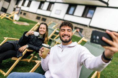 Genç bir erkek ve kadın manzaralı bir dağ evinde hafta sonu kaçamağının tadını çıkarıyorlar. Şezlonglarda uzanırken, o anı, akıllı telefonlarıyla neşeli bir selfie çekerek yakalıyorlar..