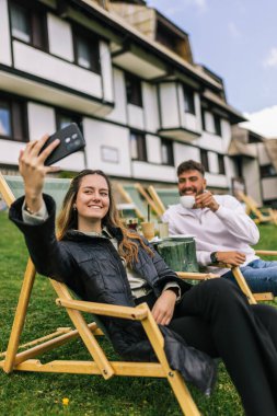 Genç bir erkek ve kadın manzaralı bir dağ evinde hafta sonu kaçamağının tadını çıkarıyorlar. Şezlonglarda uzanırken, o anı, akıllı telefonlarıyla neşeli bir selfie çekerek yakalıyorlar..
