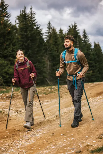 Sırt çantaları, yürüyüş direkleri ve açık hava kıyafetleriyle donatılmış genç bir erkek ve kadın, yemyeşil doğayla çevrili bir dağ yolunda birlikte yürüyorlar.