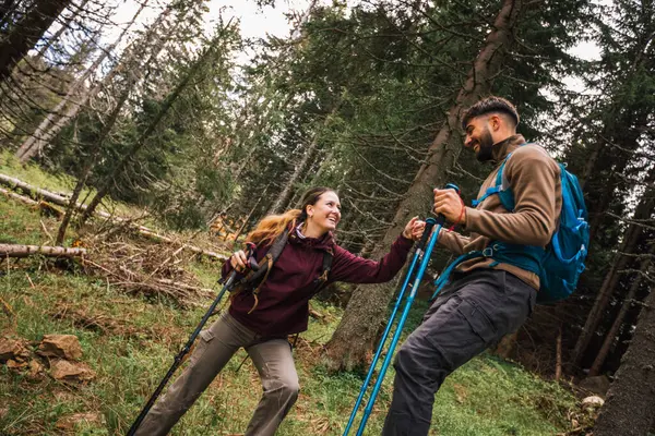 Genç bir adam, ormanlık bir dağ patikasının zorlu bir bölümünde bir kadına yardım ediyor. İkisi de tam teçhizatlı yürüyüş kıyafetleri içinde, sırt çantaları, yürüyüş kıyafetleri ve direkler..