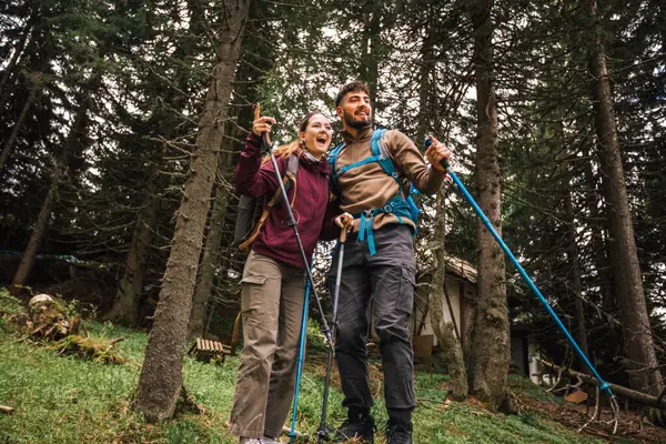 Genç bir adam ve kadın, sırt çantaları, yürüyüş direkleri ve uygun açık hava kıyafetleriyle dolu manzaralı bir dağ ormanında yürüyüş yapıyorlar. Uzaktaki bir şeye bakmak için yolda dururlar, merak ve keşif anlarını paylaşırlar..