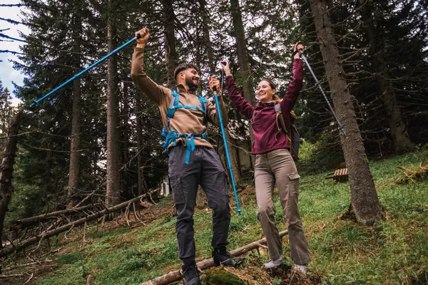 Yürüyüş kıyafetleri giymiş, sırt çantaları ve sırıklar taşıyan genç bir adam ve kadın başarılı dağ yürüyüşlerini kutluyorlar. Manzaralı bir patikada birlikte durup gülümsüyor ve neşeyi ifade ediyorlar..