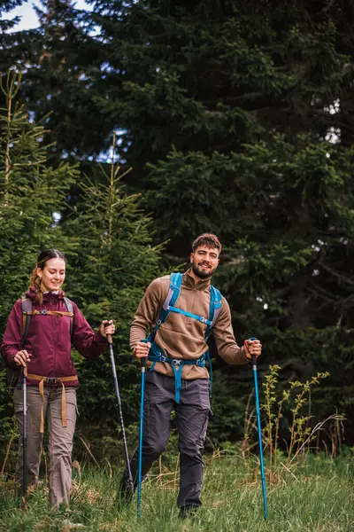 Sırt çantaları, yürüyüş direkleri ve yürüyüş elbiseleriyle donatılmış genç bir adam ve kadın yoğun çalılarla çevrili engebeli bir dağ yolunda dikkatle ilerliyorlar.. 