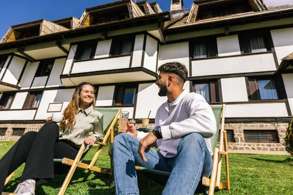 Genç bir adam ve bir kadın, hafta sonu kaçamağı sırasında şezlonglarda dinleniyorlar. Doğa ve temiz havayla çevrili olarak sıcak kahvenin ve ferahlatıcı meyve suyunun tadını çıkarırlar..