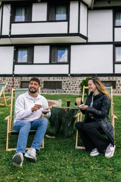 Genç bir adam ve bir kadın, hafta sonu kaçamağı sırasında şezlonglarda dinleniyorlar. Doğa ve temiz havayla çevrili olarak sıcak kahvenin ve ferahlatıcı meyve suyunun tadını çıkarırlar..