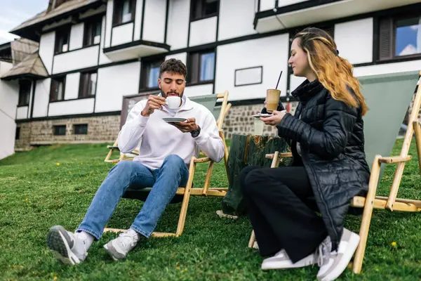 Genç bir adam ve bir kadın, hafta sonu kaçamağı sırasında şezlonglarda dinleniyorlar. Doğa ve temiz havayla çevrili olarak sıcak kahvenin ve ferahlatıcı meyve suyunun tadını çıkarırlar..