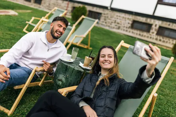 Genç bir erkek ve kadın manzaralı bir dağ evinde hafta sonu kaçamağının tadını çıkarıyorlar. Şezlonglarda uzanırken, o anı, akıllı telefonlarıyla neşeli bir selfie çekerek yakalıyorlar..