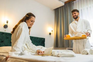 Sıcak beyaz bornozlu genç bir kadın yatağına uzanmış gülümsüyor ve erkek arkadaşı ona kahvaltı hazırlıyor..