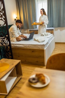 Kız arkadaşı ona kahvaltı hazırlarken, beyaz bornozlu genç adam yatakta gülümsüyor..