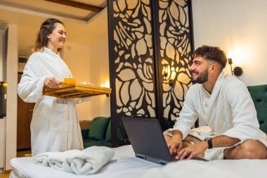 Kız arkadaşı ona kahvaltı hazırlarken, beyaz bornozlu genç adam yatakta gülümsüyor..