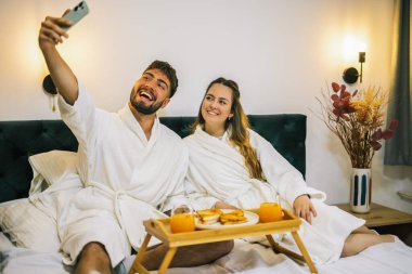 Neşeli bir çift yatakta birlikte selfie çekerek neşeli ve samimi bir anı yakalıyor. Rastgele giyinmiş ve rahat yataklarla çevrili..
