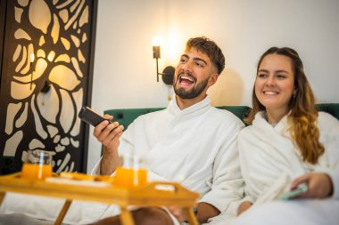 Yumuşak beyaz bornozlu bir çift TV izlerken yatakta dinleniyor ve tatillerinde huzurlu bir sabah geçiriyorlar..
