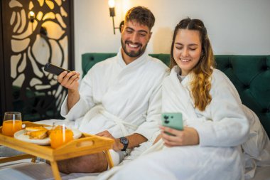 Yumuşak beyaz bornozlu bir çift TV izlerken yatakta dinleniyor ve tatillerinde huzurlu bir sabah geçiriyorlar..