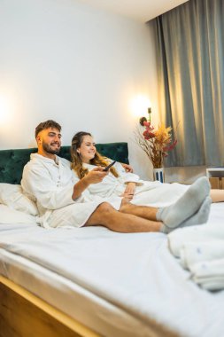 Yumuşak beyaz bornozlu bir çift TV izlerken yatakta dinleniyor ve tatillerinde huzurlu bir sabah geçiriyorlar.. 
