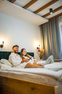 Yumuşak beyaz bornozlu bir çift TV izlerken yatakta dinleniyor ve tatillerinde huzurlu bir sabah geçiriyorlar.. 