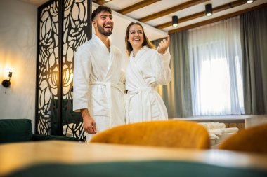 Beyaz bornoz giymiş bir çift otel odalarında yan yana durur, etrafa bakar ve içeriyi seyrederler..