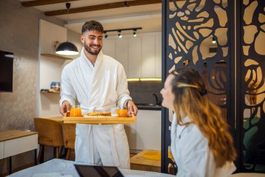 Sıcak beyaz bornozlu genç bir kadın yatağına uzanmış gülümsüyor ve erkek arkadaşı ona kahvaltı hazırlıyor..