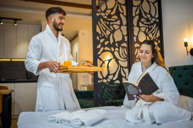 Sıcak beyaz bornozlu genç bir kadın yatağına uzanmış gülümsüyor ve erkek arkadaşı ona kahvaltı hazırlıyor..