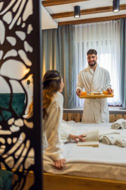 Sıcak beyaz bornozlu genç bir kadın yatağına uzanmış gülümsüyor ve erkek arkadaşı ona kahvaltı hazırlıyor..